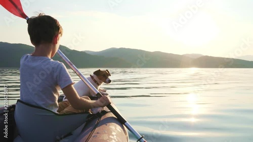 Slow motion of a happy boy with his dog Jack Russell Terrier paddling an inflatable kayak on the water of a large mountain lake against the backdrop of beautiful orange sunset. Family sports vacation