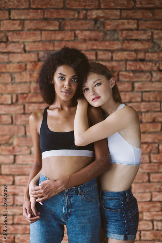 Young pretty african american woman with dark curly hair and nice woman with blond hair dreamily looking in camera together with brick wall on background isolated Young pretty african american woman with dark curly hair and nice woman with blond hair dreamily looking in camera together with brick wall on background isolated