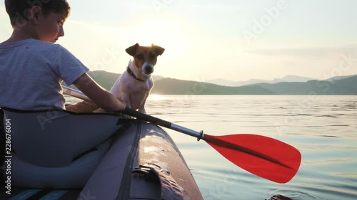 Slow motion of a happy boy with his dog Jack Russell Terrier paddling an inflatable kayak on the water of a large mountain lake against the backdrop of beautiful orange sunset. Family sports vacation