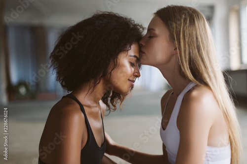 Close up young woman with blond hair thoughtfully kissing in forehead smiling african american woman with dark curly hair while spending time together at home photo