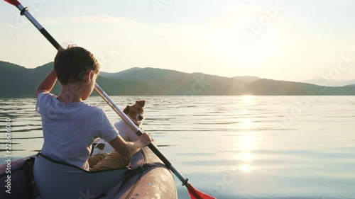Slow motion of a happy boy with his dog Jack Russell Terrier paddling an inflatable kayak on the water of a large mountain lake against the backdrop of beautiful orange sunset. Family sports vacation