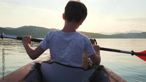 Slow motion of a happy boy with his dog Jack Russell Terrier paddling an inflatable kayak on the water mountain lake against the backdrop of beautiful orange sunset. Family sports vacation. Lens flare