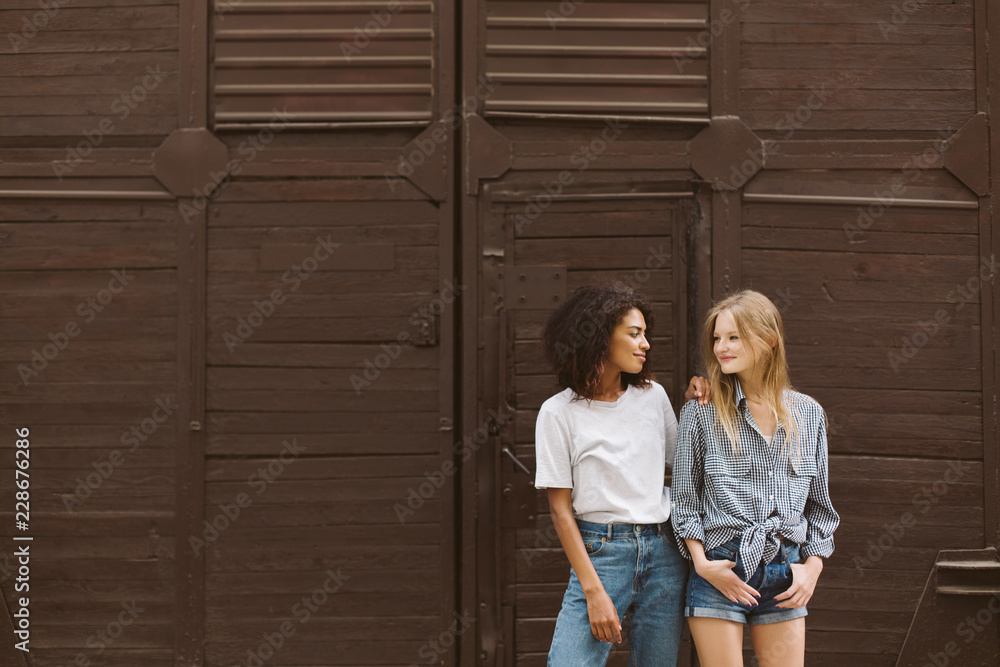 Young nice african american woman with dark curly hair in T-shirt and jeans and pretty woman with blond hair in shirt and shorts happily looking at each other with brown wall on background
