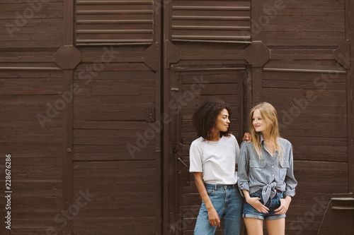 Young nice african american woman with dark curly hair in T-shirt and jeans and pretty woman with blond hair in shirt and shorts happily looking at each other with brown wall on background