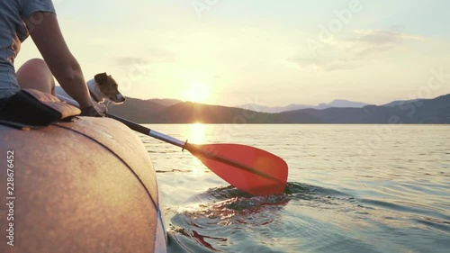 Slow motion happy young woman with her dog Jack Russell Terrier paddling on an inflatable kayak on the water of a large mountain lake against a beautiful orange sunset. Family Sports Weekend