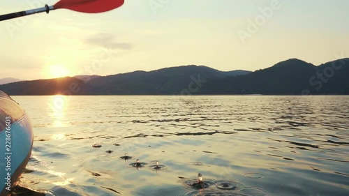 Slow motion happy young woman with her dog Jack Russell Terrier paddling on an inflatable kayak on the water of a large mountain lake against a beautiful orange sunset. Family Sports Weekend