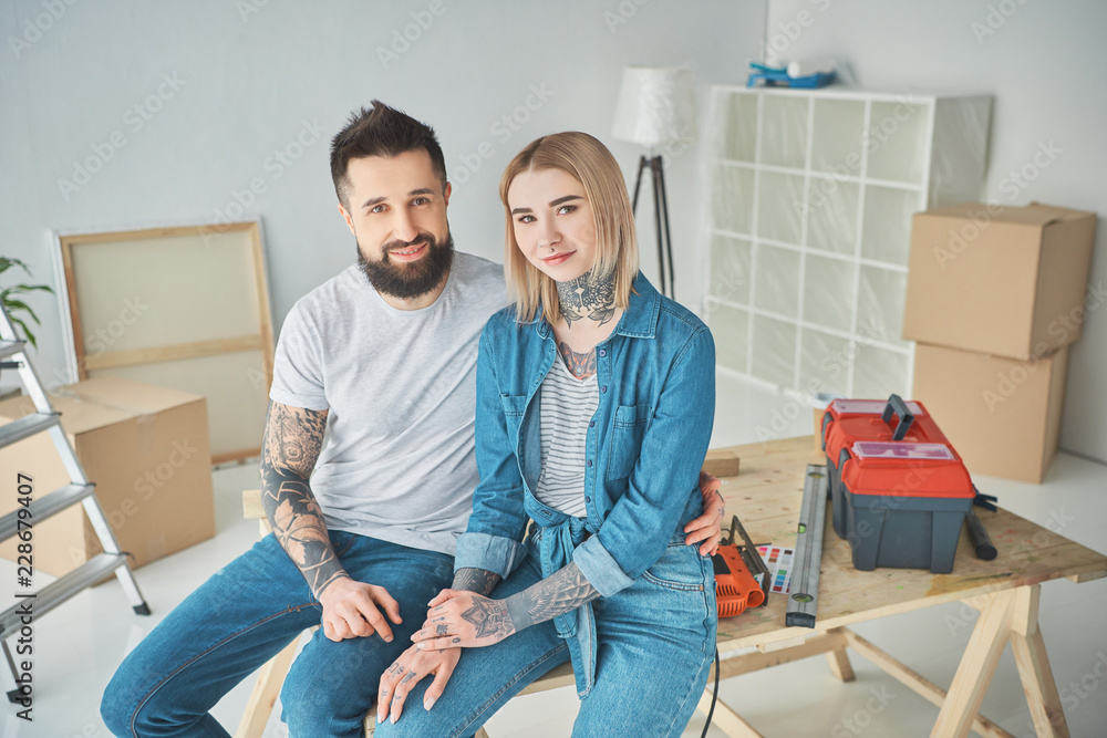 happy young couple in tattoos sitting together and smiling at camera while making repairs in new house
