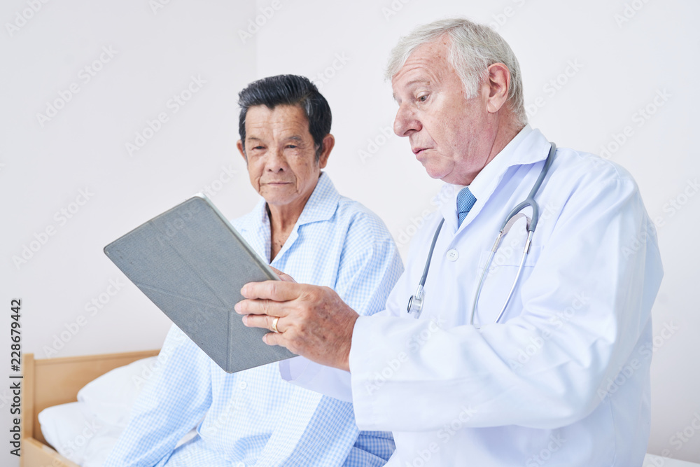 Side view of elderly medical practitioner sitting on bed near Asian male patient and showing him modern tablet Side view of elderly medical practitioner sitting on bed near Asian male patient and showing him modern tablet