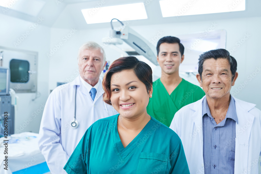 Asian woman in scrubs smiling and looking at camera while standing near male colleagues in operating theatre Asian woman in scrubs smiling and looking at camera while standing near male colleagues in operating theatre