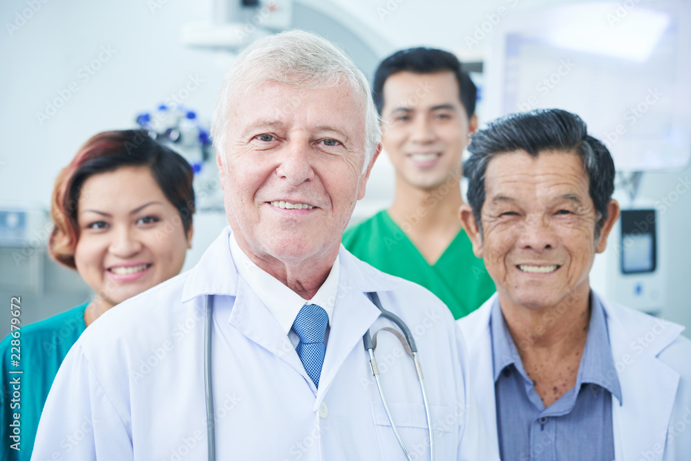 Senior medical practitioners and young Asian surgeons friendly smiling and looking at camera while standing in hospital together Senior medical practitioners and young Asian surgeons friendly smiling and looking at camera while standing in hospital together