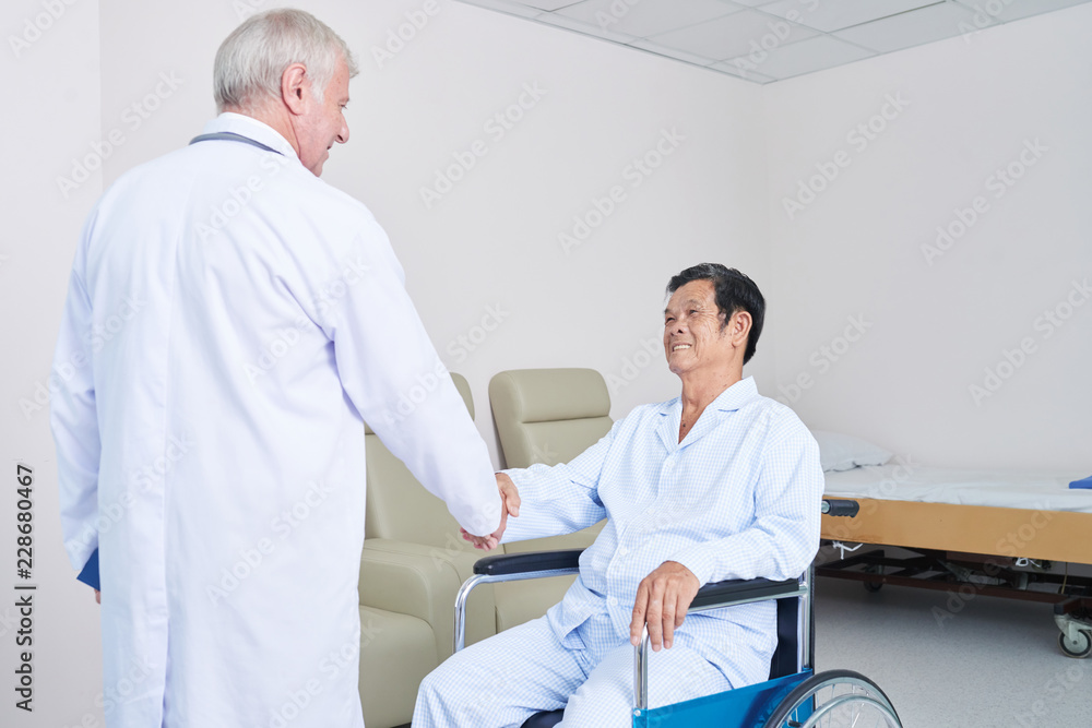 Obraz premium Elderly man in doctor uniform shaking hand of Asian male patient on wheelchair in modern ward of hospital