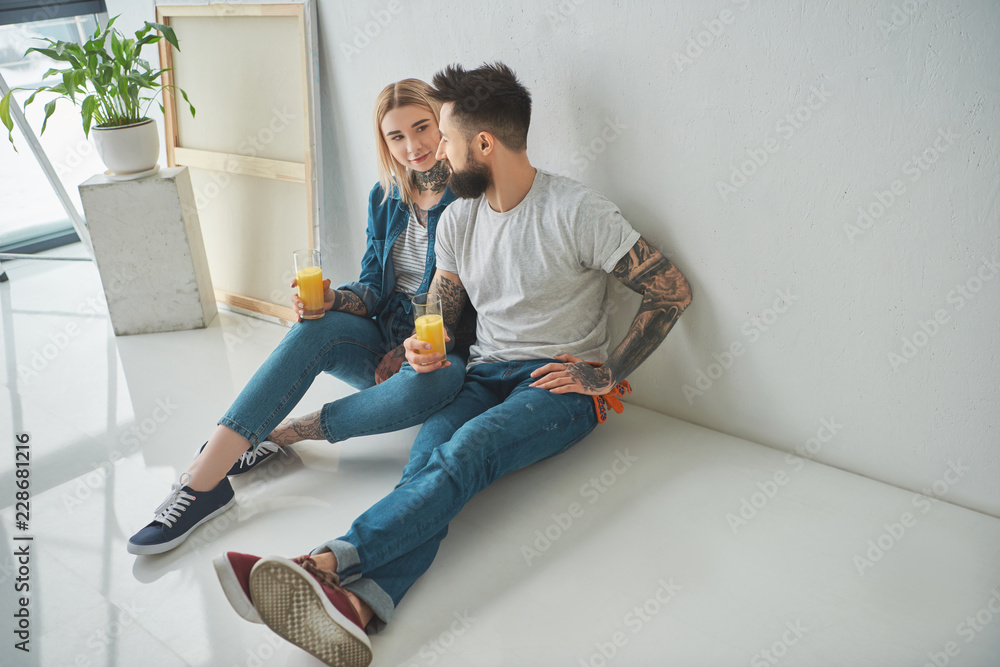 happy young tattooed couple holding glasses of juice and sitting on floor in new house