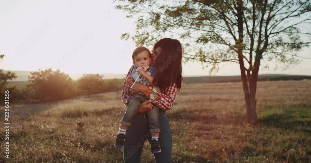 Smiling young mother with her child on the middle of field , at sunset hugging and kissing her baby , the baby eating a lollipop. Smiling young mother with her child on the middle of field , at sunset hugging and kissing her baby , the baby eating a lollipop.