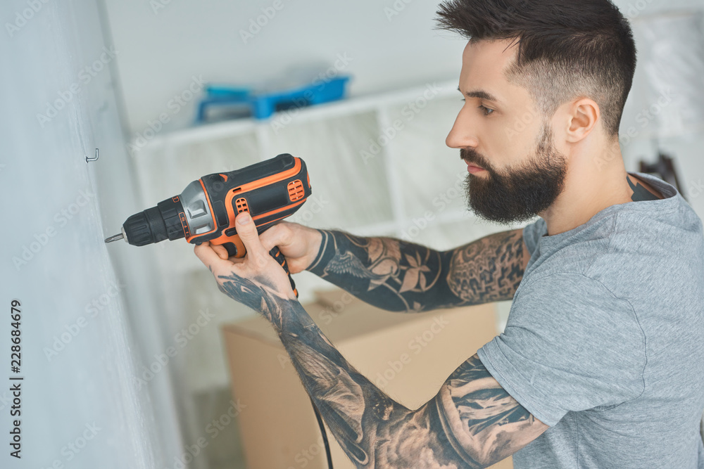 tattooed man doing hole in wall with screw gun in new apartment, inexperienced millennial concept