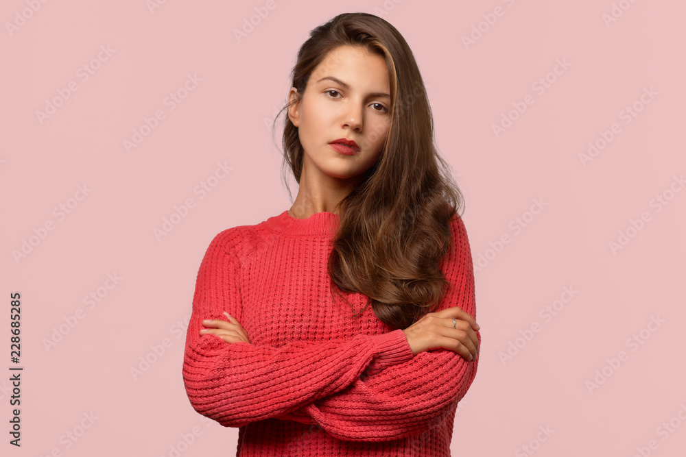 Obraz premium Beautiful long-haired brunette girl crossed her arms on her chest and looks carefully at the camera. Gaze and serious expression