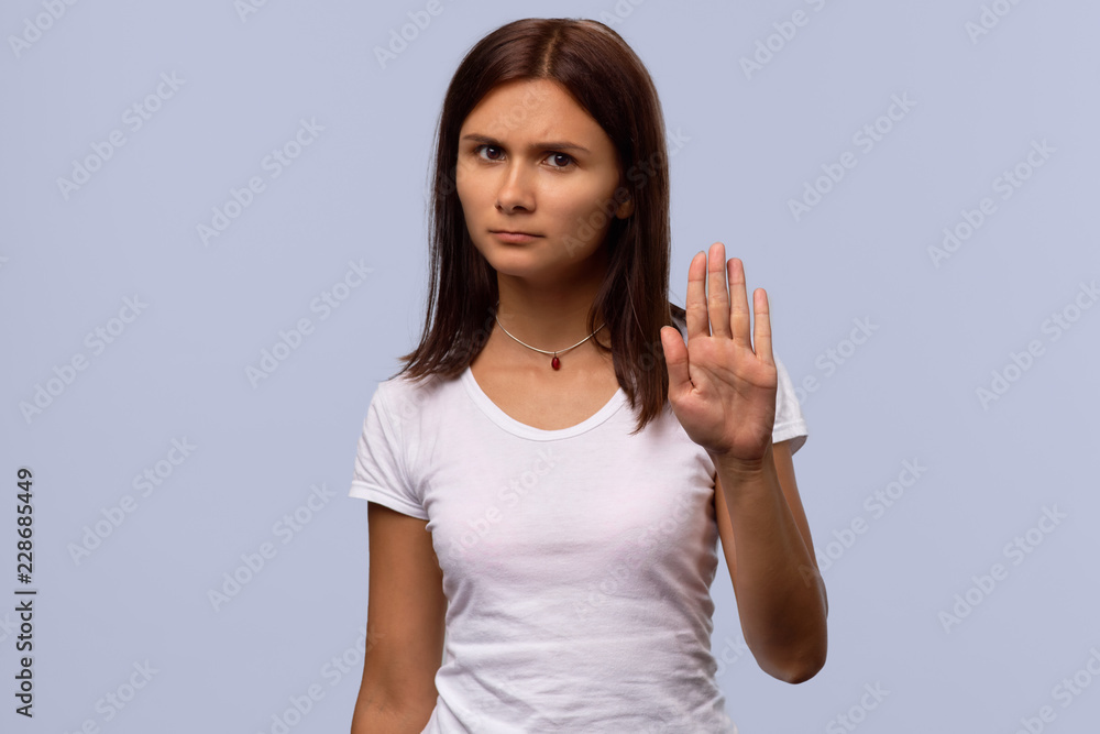Brunette girl tries to prevent herself from something unpleasant isolated on blue background. A woman in an ordinary white t-shirt shows a stop sign, prohibits doing something Brunette girl tries to prevent herself from something unpleasant isolated on blue background. A woman in an ordinary white t-shirt shows a stop sign, prohibits doing something