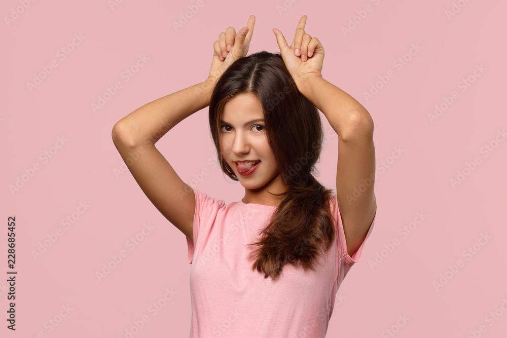 A funny young girl with long dark hair grimaces, shows her tongue and makes horns with her hands. Studio portrait on isolated pink background