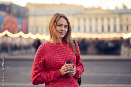 Red-haired sweet girl in a warm sweater drinking coffee while walking around the evening city in the cool autumn weather