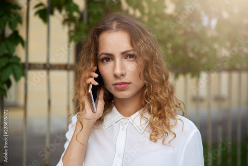A focused young girl in a white blouse looks closely at the camera while talking on the phone. Portrait of a curly-haired woman talking on the smartphone