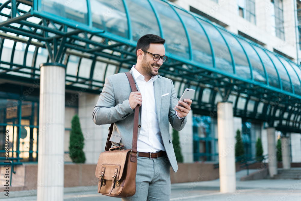 Young successful happy businessman looking at the phone and smiling. Wearing elegant suit, business bag and modern glasses, going on a business trip Young successful happy businessman looking at the phone and smiling. Wearing elegant suit, business bag and modern glasses, going on a business trip