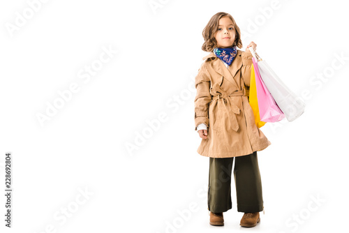 stylish little child in trench coat with colorful shopping bags isolated on white
