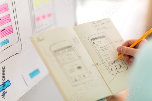 technology, user interface design and people concept - hand of ui designer or developer drawing smartphone sketches in notebook at office