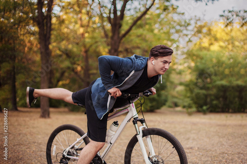 Attractive young caucasian man with dark hair bicycling in the park. White earphones, favorite music. Outdoors, golden leaves. Early autumn / fall background. Healthy life sport, fitness. Copy space