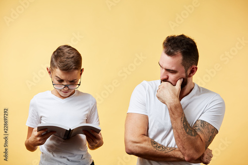 father incredulously looking at cute son reading book at yellow background photo