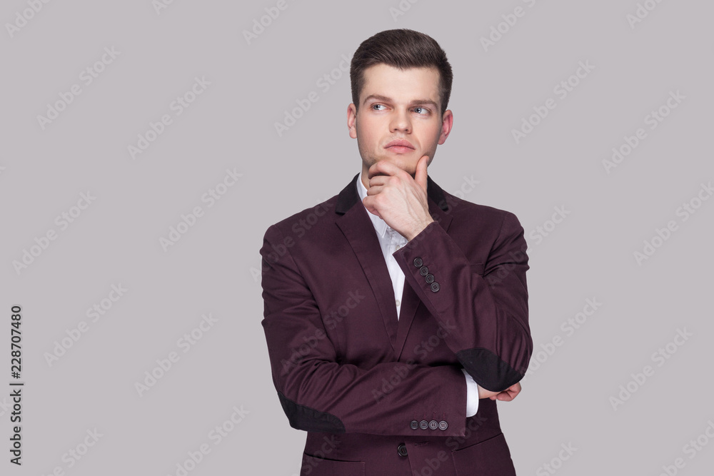 Portrait of serious thinking handsome young man in violet suit and white shirt, standing, touching his chin and looking away with thoughtful face. indoor studio shot, isolated on grey background. Portrait of serious thinking handsome young man in violet suit and white shirt, standing, touching his chin and looking away with thoughtful face. indoor studio shot, isolated on grey background.