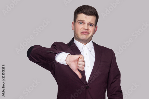 Portrait of handsome confused young man in violet suit and white shirt, standing, looking at camera with thumbs down and unsatisfied face. indoor studio shot, isolated on grey background.