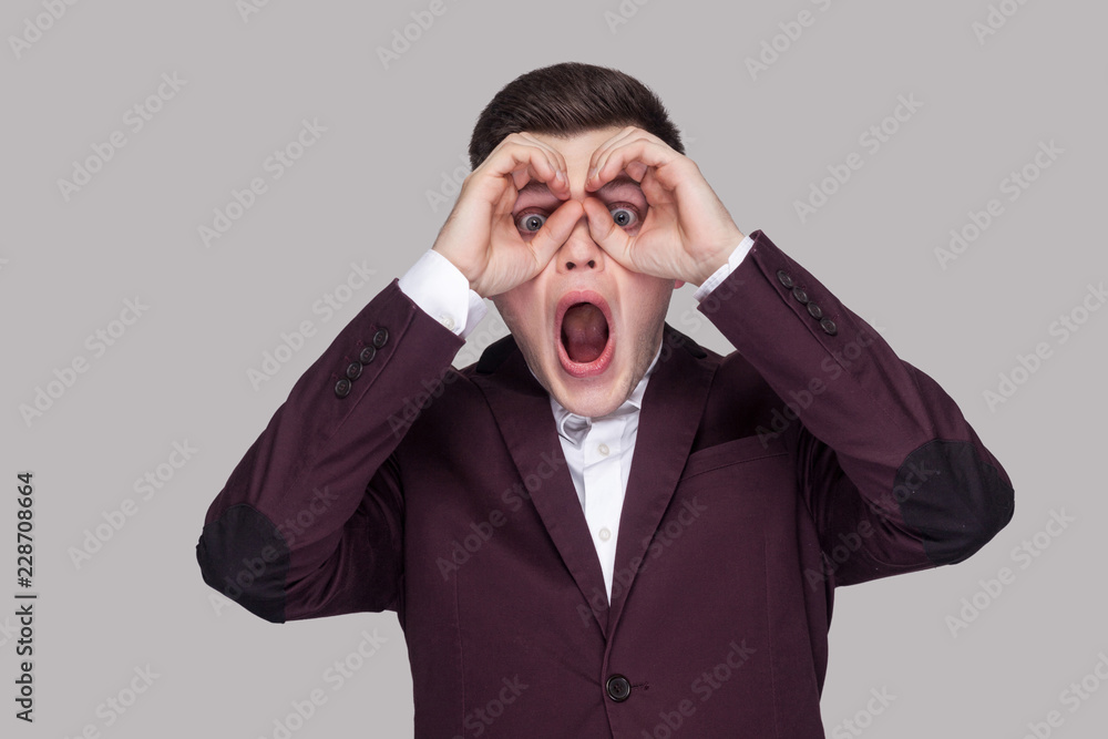 Portrait of amazed handsome young man in violet suit and white shirt, standing, looking at camera, shocked and screaming with binoculars gesture. indoor studio shot, isolated on grey background. Portrait of amazed handsome young man in violet suit and white shirt, standing, looking at camera, shocked and screaming with binoculars gesture. indoor studio shot, isolated on grey background.