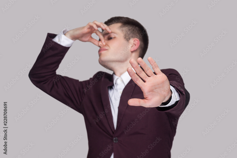 Portrait of handsome confused young man in violet suit and white shirt, standing, blocking his nose and showing stop gesture. focus on hand. indoor studio shot, isolated on grey background. Portrait of handsome confused young man in violet suit and white shirt, standing, blocking his nose and showing stop gesture. focus on hand. indoor studio shot, isolated on grey background.