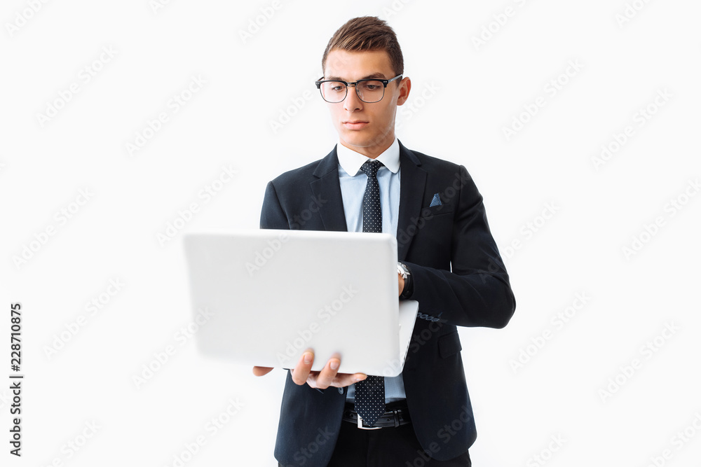 Confident man with glasses, young handsome businessman in a suit holding a laptop and looking at the screen, standing on a white background Confident man with glasses, young handsome businessman in a suit holding a laptop and looking at the screen, standing on a white background
