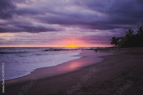 Tourists in the distance watch a pink and purple sunset over low tubular clouds on Playa Carmen in Santa Teresa, on the Pacific coast of Costa Rica