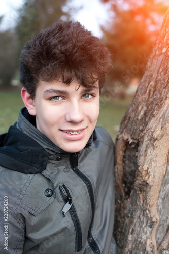 Cute, handsome teenager resting in a park leaning on a tree. The guy smiles and his braces are clearly visible on his teeth