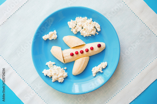 Fototapeta Fun food idea for kids. children's Breakfast: plane made of banana and clouds made of curd on a blue plate. dreams of flying. creative lunch of the future pilot