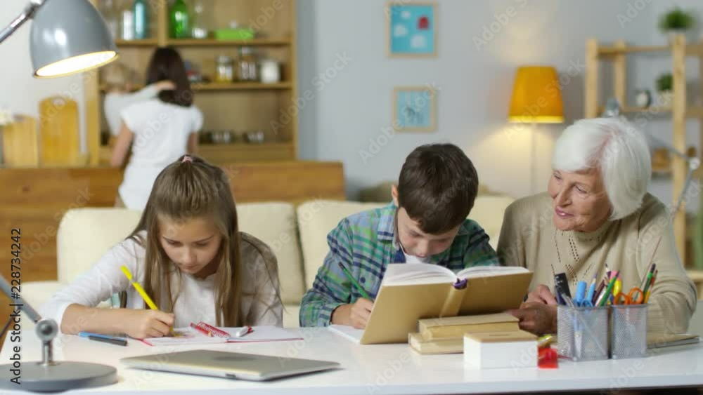 Brother and sister doing homework with help of grandmother at desk in the living room while mother walking with baby in the background Brother and sister doing homework with help of grandmother at desk in the living room while mother walking with baby in the background