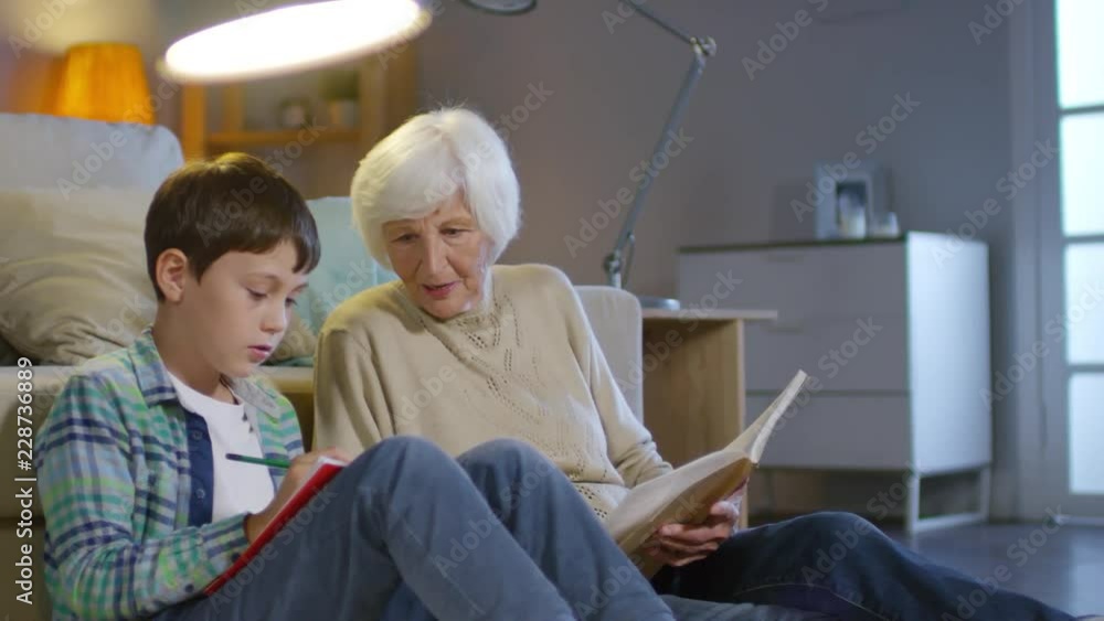 Grandmother and boy sitting on the floor by couch in the living room, talking and reading textbook while doing homework Grandmother and boy sitting on the floor by couch in the living room, talking and reading textbook while doing homework