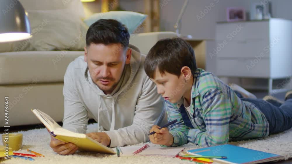 Father and son lying on comfortable carpet in the living room, reading textbook and discussing something while doing homework together Father and son lying on comfortable carpet in the living room, reading textbook and discussing something while doing homework together