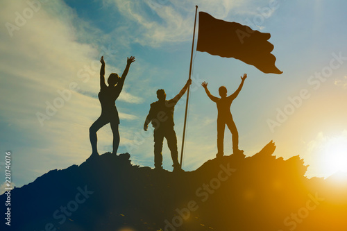 Silhouettes of happy three people on top of a mountain with the flag of victory. Against the background of the sunset. Businessmen, teamwork, winners. Success and Achievements