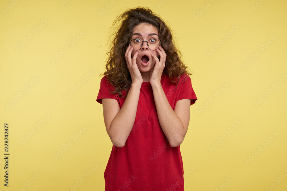 Cute brunette woman with curly hair and eyeglasses in red t-shirt, opened her mouth in a surprise, against yellow background Cute brunette woman with curly hair and eyeglasses in red t-shirt, opened her mouth in a surprise, against yellow background