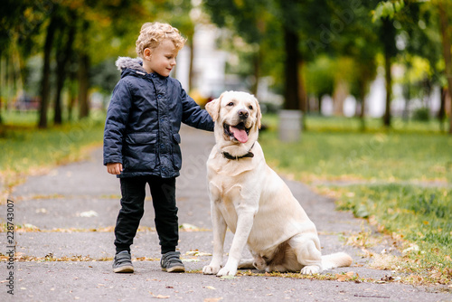Portrait of cute adorable little Caucasian baby boy sitting with dog in park outside. Smiling child holding animal domestic pet. Happy childhood concept
