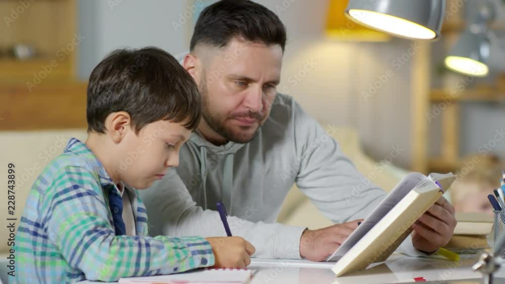 Helpful father reading textbook and talking with son while doing homework together at home Helpful father reading textbook and talking with son while doing homework together at home