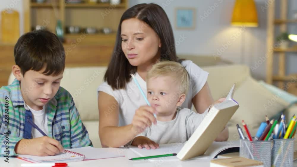 Beautiful mother helping son with homework while little baby boy sitting on her knees an looking at brother in the living room Beautiful mother helping son with homework while little baby boy sitting on her knees an looking at brother in the living room