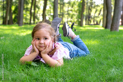 The little girl is lying on the green grass in the park and dreamily looks into the distance