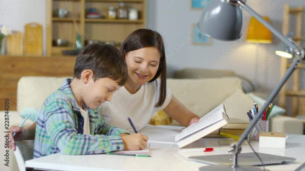 Beautiful mother smiling and talking to son while doing homework together at desk in the living room Beautiful mother smiling and talking to son while doing homework together at desk in the living room