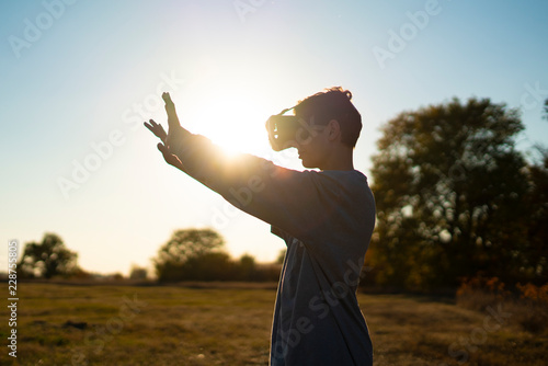 young man in VR glasses play games outdoors in the park during sunset against the sky а