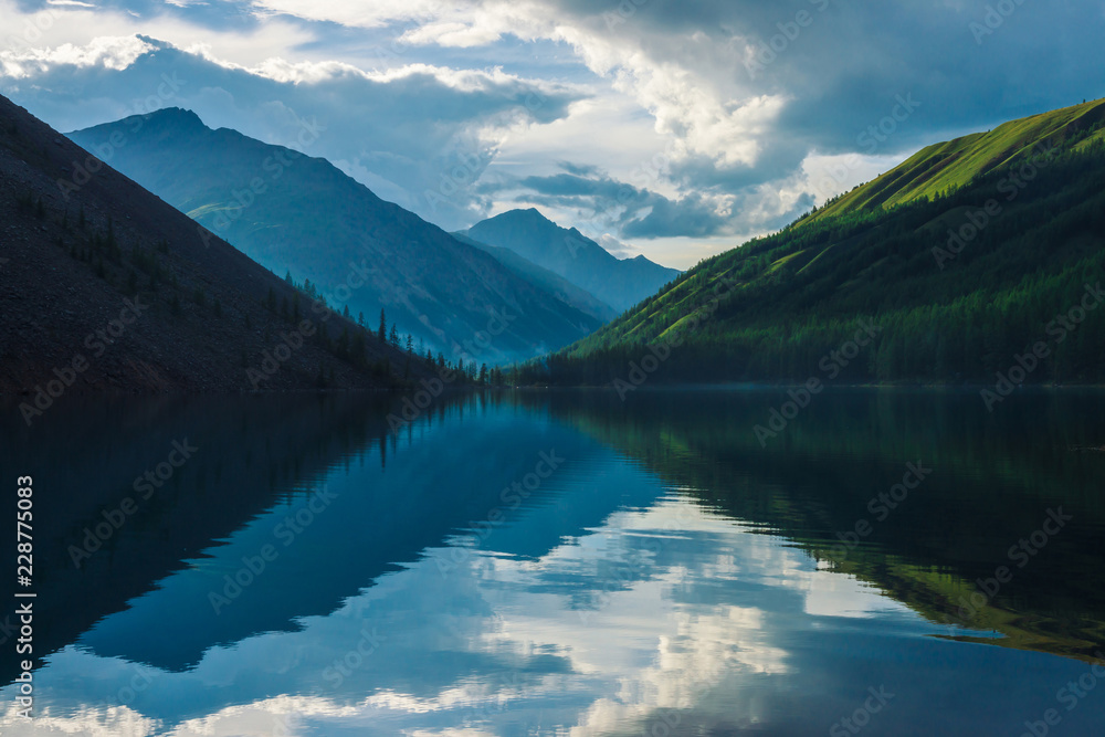 Ghostly mountain lake in highlands at early morning. Beautiful misty silhouettes of mountains and clouds reflected in clear water surface. Smoke of campfires. Amazing landscape of majestic nature.