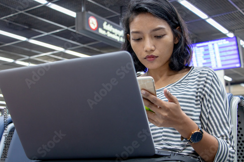 A young woman with mobile phone in her hand is working on a computer in an airport lobby.