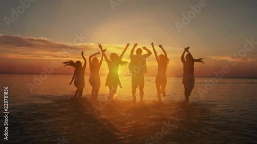 Big crowd of friends having fun at sunset sea beach. Beach vacations concept. Beauty and joyful teenager friends having fun, dancing, spraying over summer sunset. Beach party. Sun flare. Slow motion